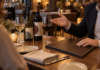 Eine Person gestikuliert an einem Restauranttisch neben zwei Weinflaschen und Weingläsern; davor liegt eine geschlossene Mappe.
