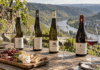 Vier deutsche Weinflaschen und gefüllte Gläser vor einer Weinlandschaft mit Flusstal.